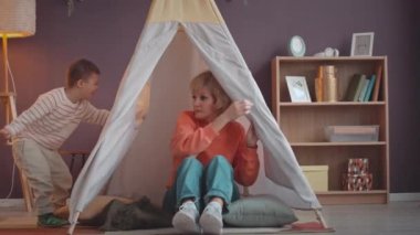Down sendromlu neşeli beyaz bir çocuk ve sevgili annesi boş vakitlerini birlikte rahat bir oyun çadırında parlak, ferah oturma odasında oturarak geçiriyorlar.