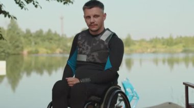 Tekerlekli sandalyede dalgıç kıyafetli beyaz adamın orta boy portresi. Nehrin kıyısındaki ahşap rıhtımda kameraya bakıyor.