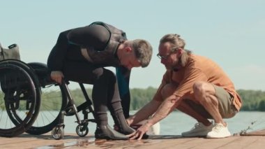 Engelli bir adamın tekerlekli sandalyeden kurtulup tahta iskeleye oturmasına ve suya girmeden önce hazırlık yapmasına yardımcı olan uyarlanabilir sörf eğitmeninin yan görüntüsü.
