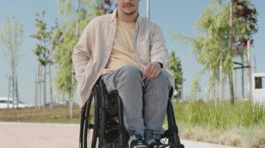 Genç beyaz adamın portresini kaldır gözlüğünü çıkar ve güneşli bir günde tekerlekli sandalyede otururken kameraya gülümse.