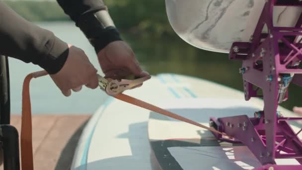 水上に行く前に彼の適応性のスーパーボード上の安全シートベルトを点検するウェットスーツの認識できない車輪修理された男のクロップショット — ストック動画