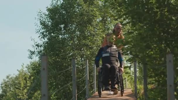 晴れた日に川沿いの木製の橋に沿って男性の友人によって車椅子で押されている障害ウェットスーツを身に着けている白人男性の全長ショット — ストック動画