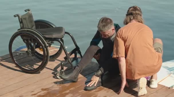 年轻的白人残疾人和他的适应性冲浪教练在阳光照射下坐在木制码头上做最后的准备 然后坐上舷梯 — 图库视频影像