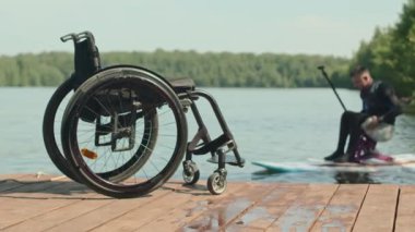 Dalgıç kıyafetiyle engelli genç atletik adam nehirde kürek çekiyor. Tahta iskelede güneş ışığı altında tekerlekli sandalyeye odaklanıyor.