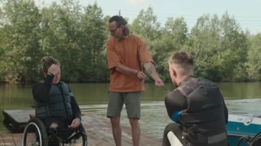 Profesyonel erkek adaptif wakeboard eğitmeni. Tahta iskelede dalgıç kıyafetli genç ve tekerlekli sandalyeli adama ders veriyor.