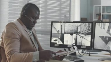 Afrika kökenli Amerikalı erkek güvenlik görevlisinin, bilgisayarın başında otururken, masaüstünde birden fazla CCTV güvenlik kamerası olan belgeleri incelemesini yavaşlat.