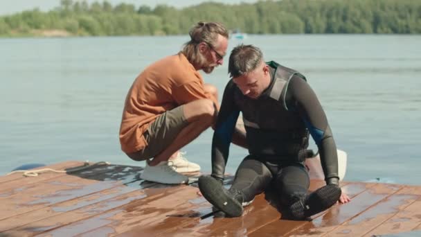 身穿残疾泳衣的白人男子和他的适应性冲浪教练坐在潮湿的木制码头上晒太阳 准备下水 — 图库视频影像