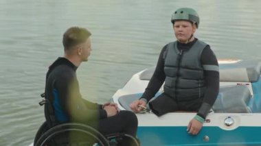 Tekerlekli sandalda oturan adaptif wakeboard eğitmeni, suda bacakları olmayan beyaz ergen çocuğa talimatlar veriyor.
