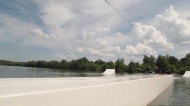 Beyaz tenli genç sörfçü dalgıç kıyafeti ve kask takmış ve kayganlaştırıcı üzerinde kayarken açık hava antrenmanı sırasında oturarak uyumlu bir wakeboard kullanıyor.