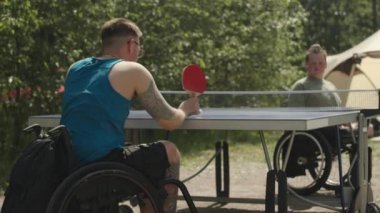 Sıcak ve güneşli bir günde dışarıda masa tenisi oynayan iki tekerlekli sandalyeli erkek arkadaşın orta yavaşlığı.