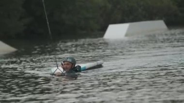 Dalgıç kıyafetli ve kasklı beyaz bir genç nehirde yüzerken wakeboard ve halata tutunarak su sporları yapıyor.