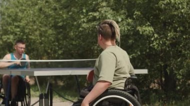 İki beyaz tekerlekli sandalyeli erkek arkadaş yazın dışarıda masa tenisi oynuyorlar.