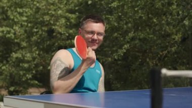 Engelli genç beyaz adam dışarıda masa tenisi oynayarak vakit geçiriyor.