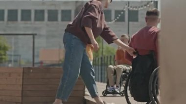 Genç beyaz kadın, yaz zamanı arkadaşlarıyla dışarıda buluşurken ahşap şehir köprüsüne çıkmak için tekerlekli sandalyeli erkek arkadaşına yardım ediyor.