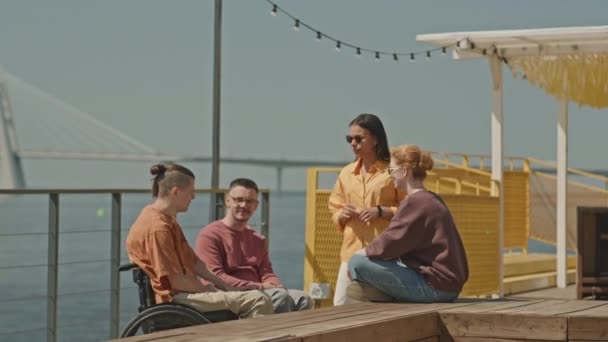 美しい川の景色で夏の遊歩道で会話しているガールフレンドと2人の若い白人車椅子の男性の中型ショット — ストック動画