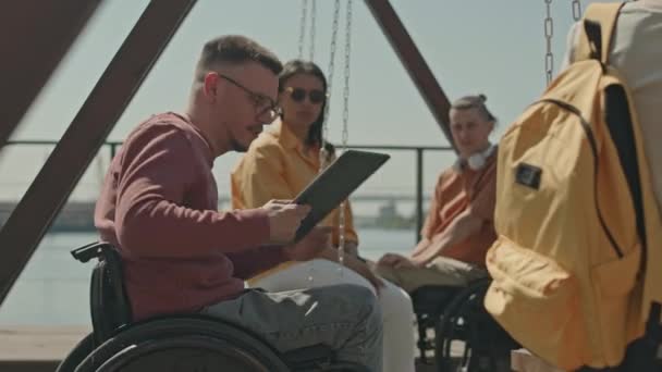 夏季与朋友在河堤散步时 坐在轮椅上的白人年轻人在户外使用笔记本电脑的中景照片 — 图库视频影像