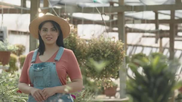 在阳光灿烂的日子里 穿着草帽 头戴棉布工作服 在大苗圃摆出一副相机的年轻美丽的中东女人的中等肖像 — 图库视频影像