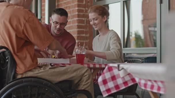 昼間は屋外サマーカフェでピザとチャットを食べる車椅子の若者とその友人のミディアムショット — ストック動画