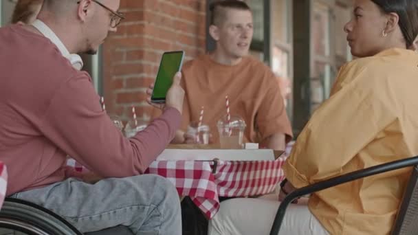 白色轮椅上的年轻人和朋友坐在外面的咖啡桌旁 一边看着智能手机上的绿色彩色按键屏幕 一边拍着照片 — 图库视频影像