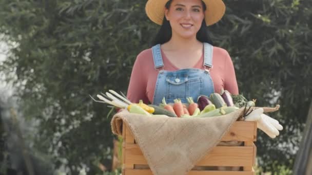 デニムカバーを身に着けている若い中東の女性農民の笑顔と 夏には屋外で新鮮な有機野菜の木箱でカメラのためにポーズするわら帽子のミディアム肖像画 — ストック動画