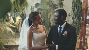 Orta halli, iyi giyimli Afro-Amerikan çiftin canlı yeşil bitkilerle dolu düğün törenlerinin güzel yerinde dikilirken sohbet edişlerinin fotoğrafı.