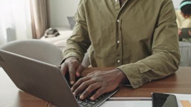 Afro-Amerikalı bir adamın portresini dizüstü bilgisayarda yazarken ve oturma odasında tahta masada otururken kameraya gülümserken arka planda tablet bilgisayarında oynayan bir çocuğun portresini kaldır.