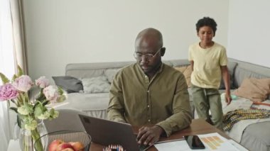 Afro-Amerikalı çocuğun orta boy portresi ve babasının evden bilgisayara poz vererek ışık minimalist oturma odasında çalışması.