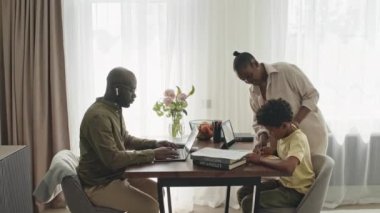 Siyah kadının, çocuk yaşta çocuğa ev ödevinde yardım ederken kocasının hafta sonları evde birlikte çalıştığı orta boy bir fotoğraf.