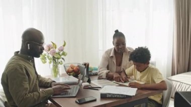 Üç kişilik modern siyahi ailenin hafta sonları evde geçirdiği orta boy bir fotoğraf. Karısı ve torunu birlikte ödev yaparken uzaktan bilgisayarla çalışan bir adam.