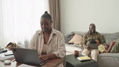 Kocası ve küçük oğlu arka planda kanepede dinlenirken, genç Afrikalı Amerikalı bir kadının laptopta çalışırken orta boy fotoğrafı.