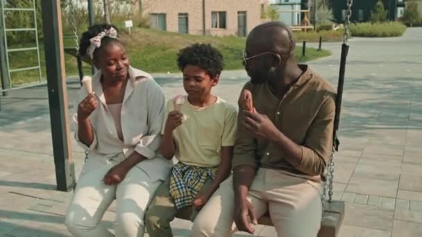 暑い夏の日に家の外で遊び場で泳いで座っている3人のチャットとアイスクリームを食べる現代アフリカ系アメリカ人の家族のミディアムショット — ストック動画