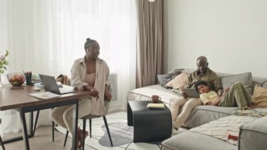 Modern, aydınlık, rahat bir dairede üç haftasonunu birlikte geçiren modern Afro-Amerikan ailesi. Kadın dizüstü bilgisayarda çalışırken, erkek ve oğlu dijital tabletle kanepede dinleniyor.