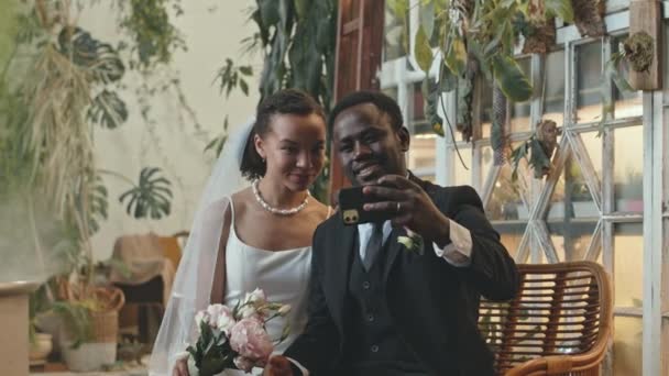 結婚式の後にスマートフォンでセルフィーの肖像画を撮る熱帯植物でいっぱいの部屋に座っている喜びの若いアフリカ系アメリカ人のカップルのミディアムショット — ストック動画