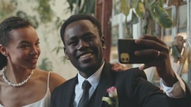 Mutlu, genç, sevecen Afrikalı Amerikalı çift düğün töreninden hemen sonra kapalı alandaki tropik bitkilerle akıllı telefondan selfie çekiyorlar.