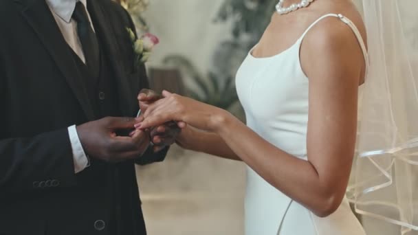 屋内での伝統的な式典の間に彼の最愛の花嫁の指に結婚指輪を置く古典的な黒いスーツの認識できない黒いグロムのクロップショット — ストック動画
