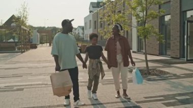 Güneşli bir günde birlikte alışveriş yaptıktan sonra sokakta yürürken ellerinde poşetlerle konuşan üç kişilik neşeli Afro-Amerikan aile fotoğrafı.