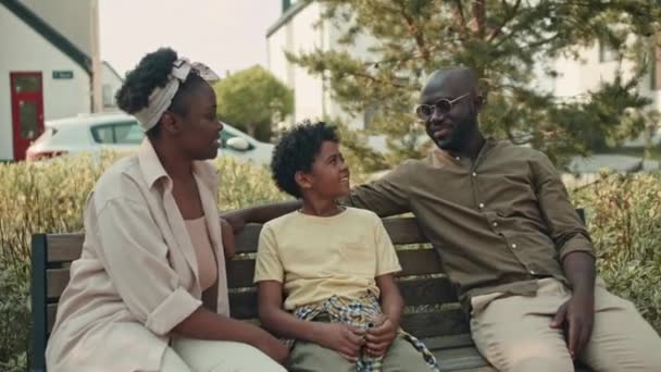家近くの広場に3人のアフリカ系アメリカ人家族が木製のベンチに座って 夏の日に一緒に楽しい時間を過ごした中型ショット — ストック動画