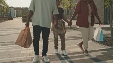 Sıcak bir yaz günü, ailesi kağıt alışveriş torbaları taşıyan mutlu Afrika kökenli Amerikalı çocuk şehir dışında kaldırımda yürürken konuşuyor.