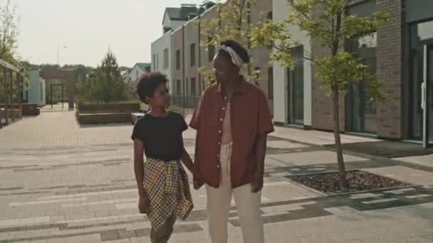 在温暖的阳光明媚的日子里 一个快乐的年轻黑人妇女和她十几岁的儿子手牵着手在户外散步聊天 — 图库视频影像