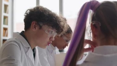 Beyaz laboratuvar önlüklü ve güvenlik gözlükleri takmış kimya dersinde konuşan bir sürü genç öğrenci.