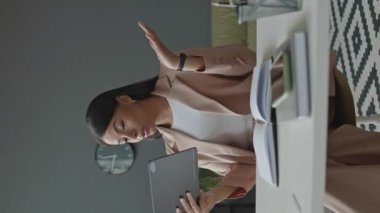 Minimalist bir ofiste çalışırken şık bir forma içinde dijital tablet üzerinde konferans veren kendine güvenen genç bir kadının dikey orta pozu.