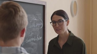 Genç beyaz kadın programcılık öğretmeni kara tahtada öğrencilere yardım ediyor, sorunları tartışıyorlar ve çözüyorlar.