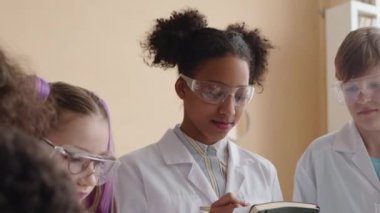 Afrika kökenli Amerikalı liseli kızın güvenlik gözlüğü ve beyaz laboratuvar önlüğü takıp okul laboratuarında sınıf arkadaşlarıyla kimya çalışırken not aldığı belden yukarı pozlar.