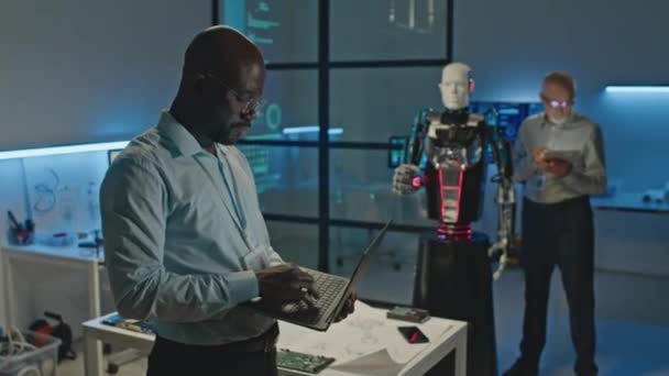 Μεσαία Λήψη Αφροαμερικανού Μηχανικού Ρομποτικής Χρήση Φορητού Υπολογιστή Ενώ Συνεργάζεται — Αρχείο Βίντεο