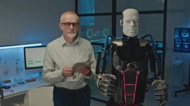 Dijital tabletli orta yaşlı beyaz erkek teknoloji geliştiricisinin orta boy portresi ve fütüristik laboratuvarda kameraya poz veren insansı robot.