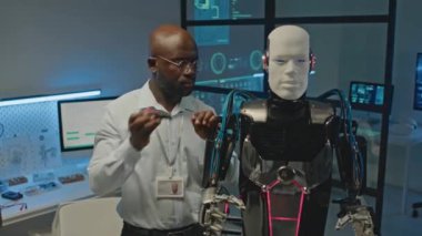 Afrika kökenli Amerikalı erkek robotik mühendisinin orta boy portresi ve fütüristik atölyede kameraya poz veren insan benzeri robot.