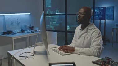 Beyaz laboratuvar önlüklü Afrika kökenli Amerikalı erkek elektronik mühendisi, karanlık modern laboratuarda bilgisayar üzerinde çalışırken beyaz masada oturuyor.