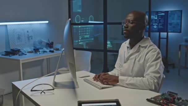アフリカ系アメリカ人の男性エレクトロニクスエンジニアが白い実験室のコートを白い机に座ってダークモダンなラボでコンピュータで作業するのを待つ — ストック動画