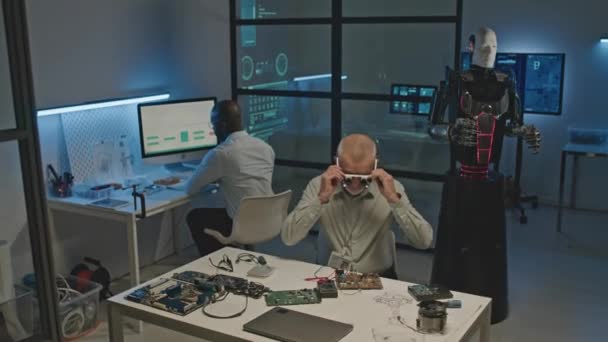 暗未来实验室工作台上聚焦成熟的高加索机器人工程师焊接线路板的中景拍摄 与同事共同开发人形机器人 — 图库视频影像