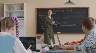Modern bilişim sınıfında karatahta ile konuşan kadın öğretmeni dinlerken program çalışması yapan çok ırklı bir grup genç öğrenci.